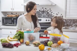 A young mother puts a sandwich in a transparent food storage bag.  His daughter hands him a peach.  They are in a bright, modern kitchen with a concrete counter surrounded by other fruits and vegetables that she prepares and stores.