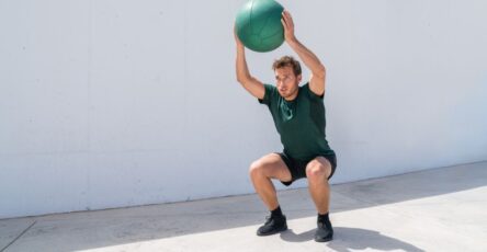 Man against a concrete background holding a medicine ball above his head in an overhead squat