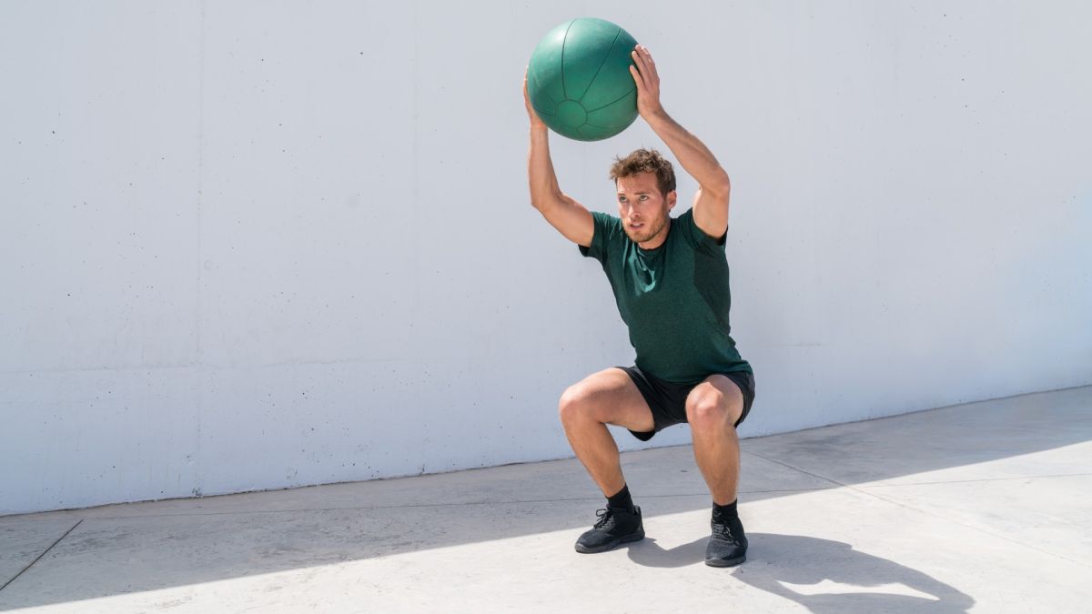 Man against a concrete background holding a medicine ball above his head in an overhead squat