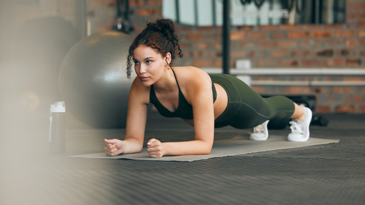 A woman does a plank on a mat in a gym.